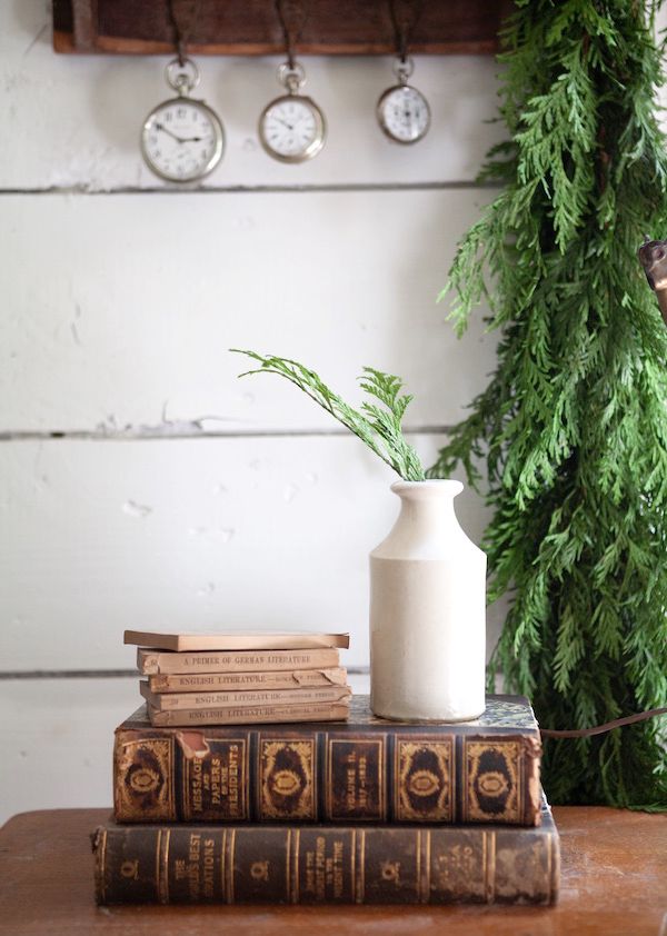 Sprig of greenery in vintage white jar sitting on old books with green garland as backdrop.