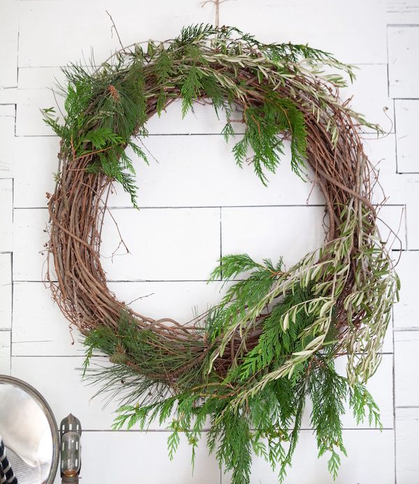 Natural wreath made from grapevine embellished with greenery hanging on white wood wall.